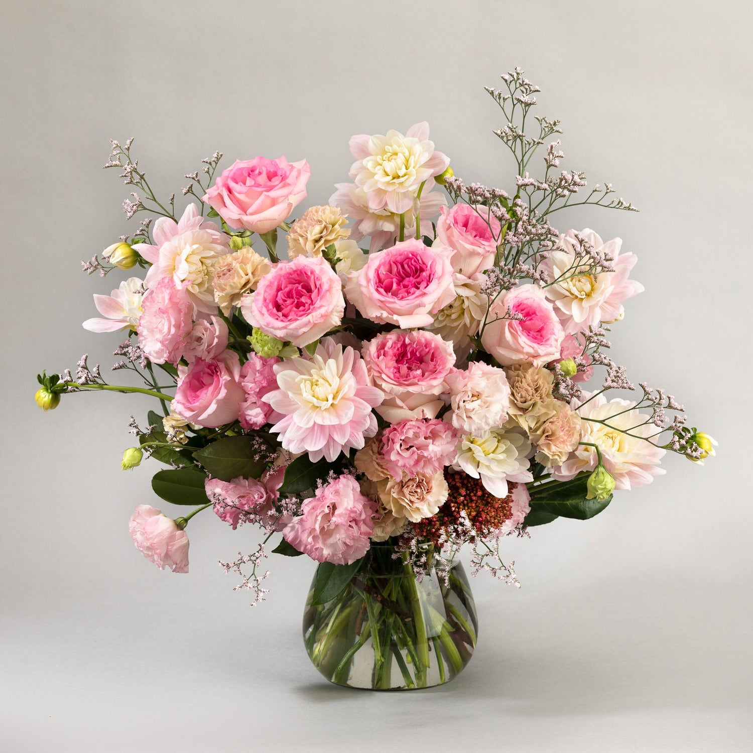 A glass vase filled with a mix of flowers in soft and bright pink. The flowers are roses, lisianthus and dahlias.