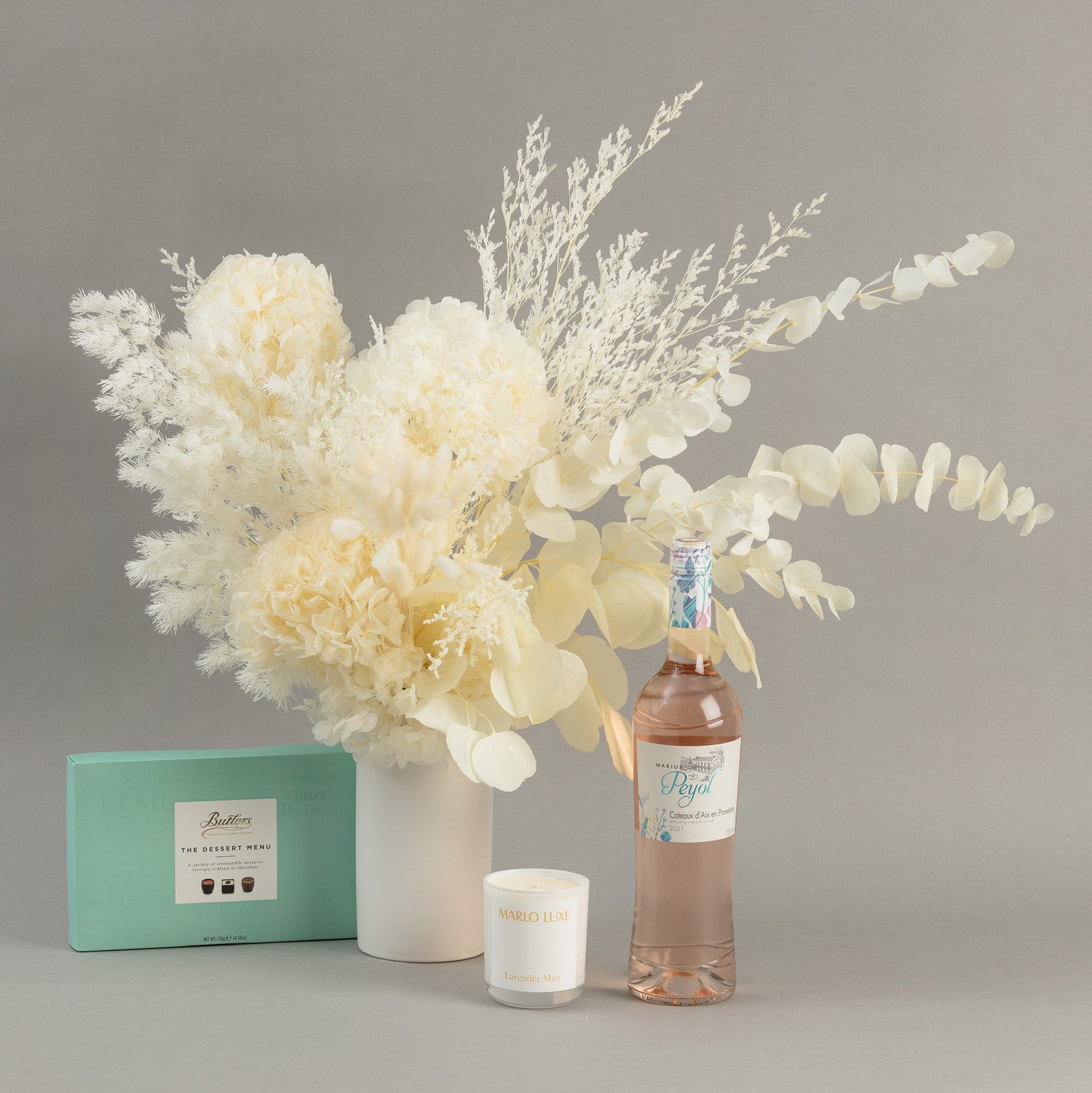 A gift package including a white ceramic vase with modern preserved flowers in cream colours, a box of premium chocolates, a bottle of wine and a candle.
