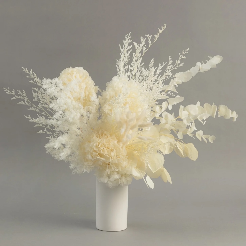 Elegant preserved floral arrangement in white tones, featuring hydrangeas, eucalyptus, and delicate foliage in a sleek white vase.